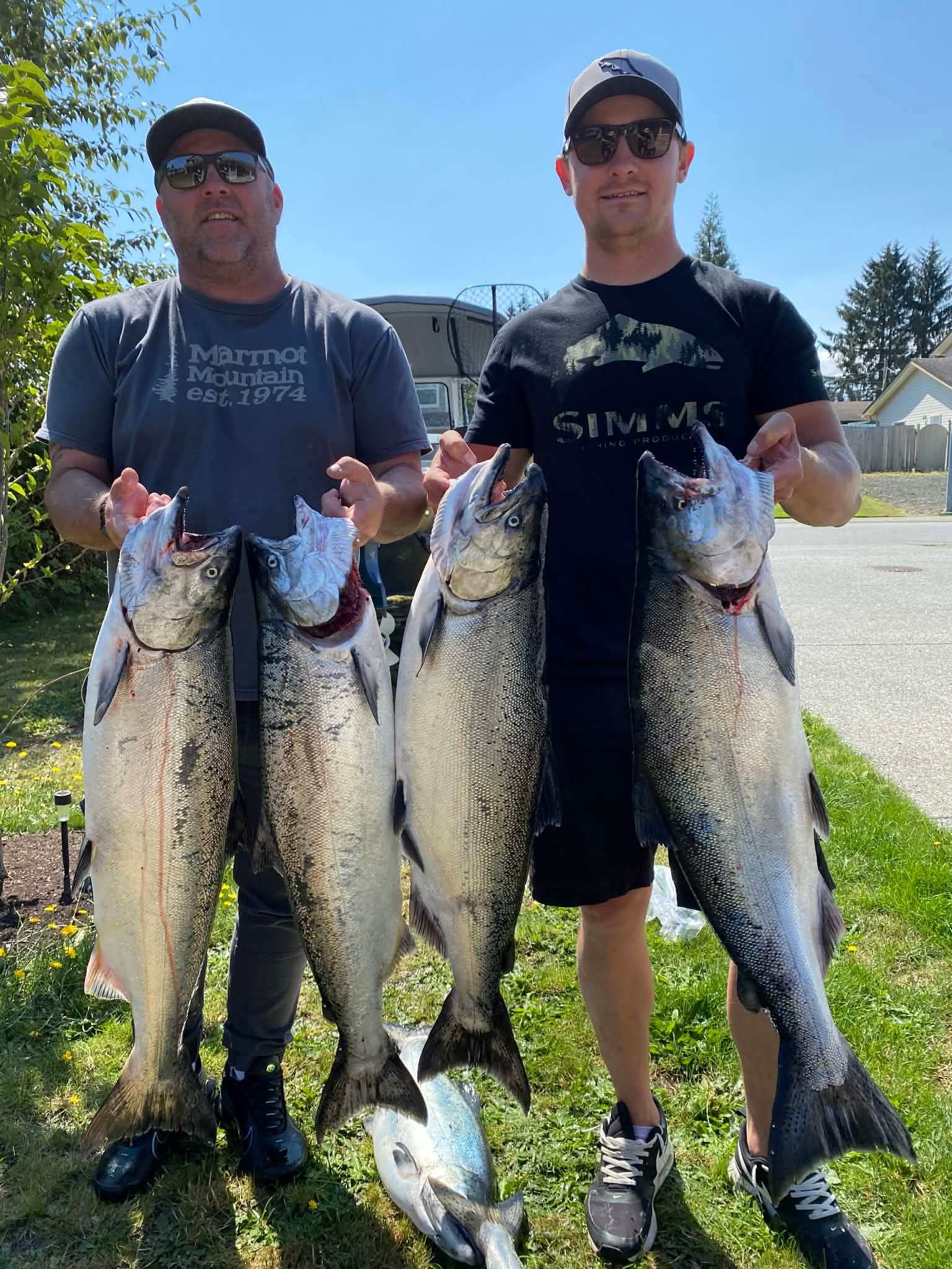 Two men smile and pose with 6 large fish they caught, the fish are silver in color and a small amount of blood is visible on the fish.
